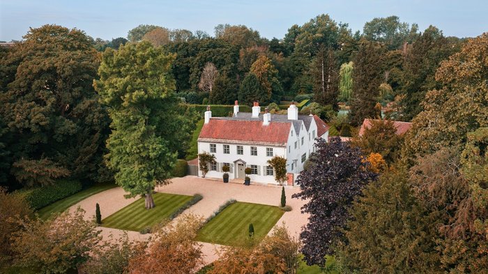 Aerial view of the white country Omaze house with a red-tiled roof, surrounded by mature trees, landscaped lawns, and gravel paths in an expansive garden.