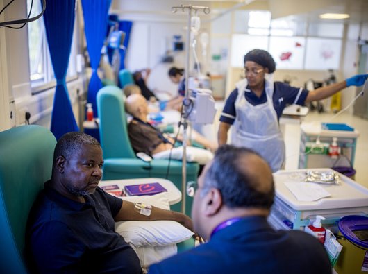 A male patient is sitting in a chair talking to a male doctor who is facing away from the camera. A female nurse is nearby, preparing a medication.