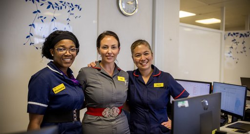 Three nurses link arms and smile to the camera. They are in a hospital.