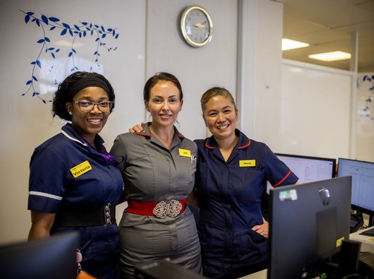 Three nurses link arms and smile to the camera. They are in a hospital.