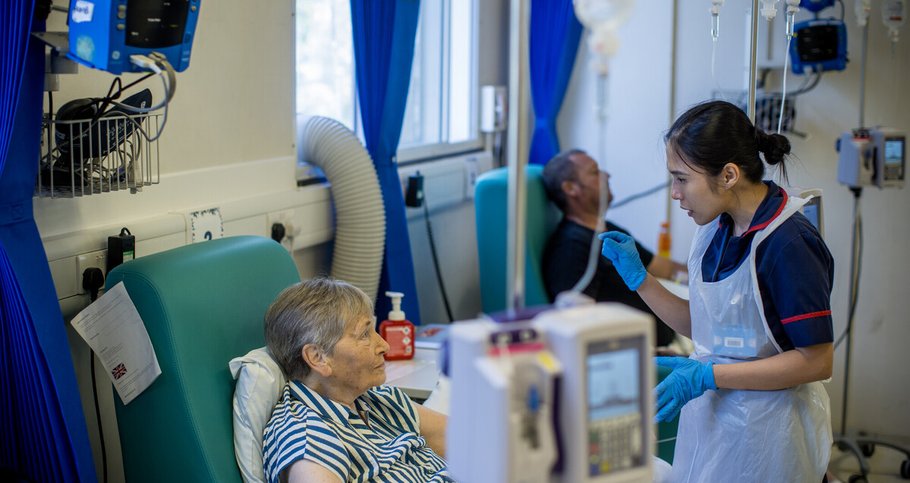 A female nurse and female patient are talking in a day unit. The patient sits in a chair, while the nurse stands chatting to her. She wears a gown and gloves. There is an infusion pump in the forefront of the picture.