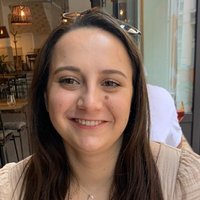 A person smiling at the camera while sitting in a café, with tables, chairs, and indoor lighting in the background.