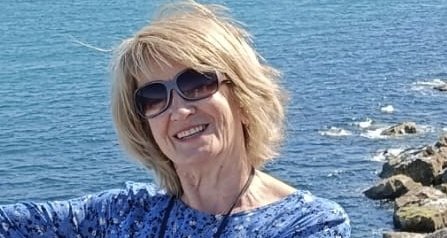 Janette is pictured smiling to the camera while wearing sunglasses. She is in front of the sea.