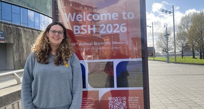 Jess, a young woman with long brown hair and glasses, standing next to a sign advertising the British Society of Haematology Scientific Congress