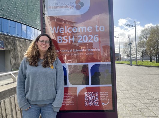 Jess, a young woman with long brown hair and glasses, standing next to a sign advertising the British Society of Haematology Scientific Congress