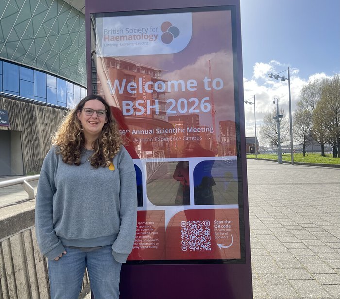 Jess, a young woman with long brown hair and glasses, standing next to a sign advertising the British Society of Haematology Scientific Congress