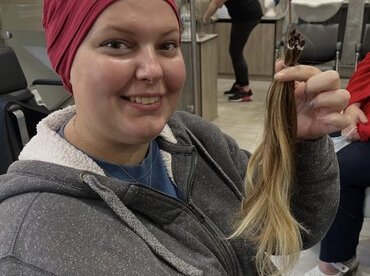 Jess sitting in a hairdresser's chair after having her hair cut off. She's wearing a red head-covering and is holding bunches of the cut hair and smiling at the camera.