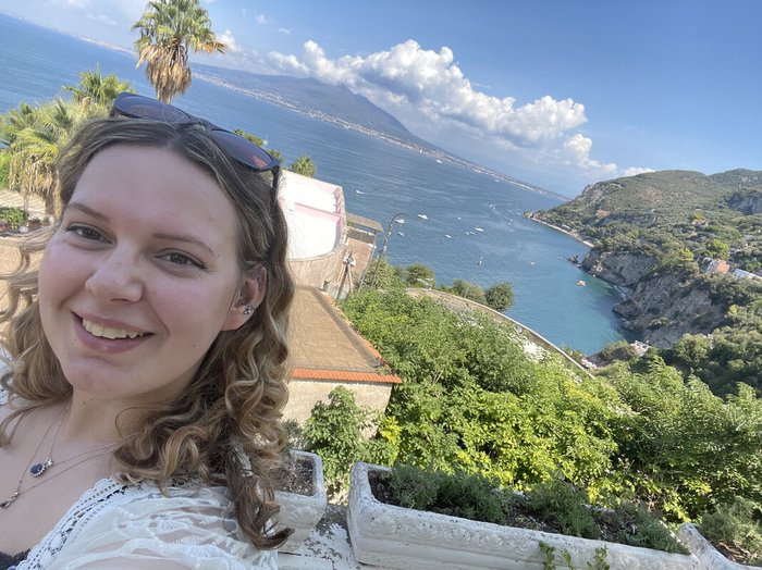 Jess on holiday, smiling at the camera with a view of blue sea and coastline in the background