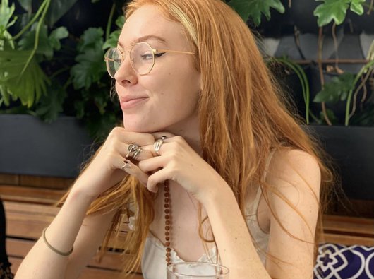 A woman in her 20s wearing glasses and with long hair smiles as she enjoys a glass of water in a cafe.
