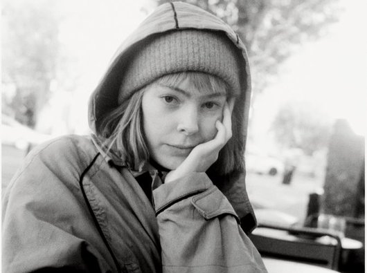 A woman in her 20s poses for a photograph outside. She's wearing a wooly hat and coat and sits at a cafe.
