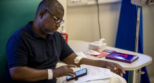 A man in hospital sits in a chair while reading his phone.