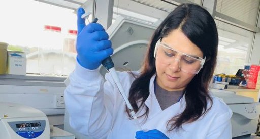 A lady working in the lab wearing lab goggles and a white lab coat, holding a pipette.