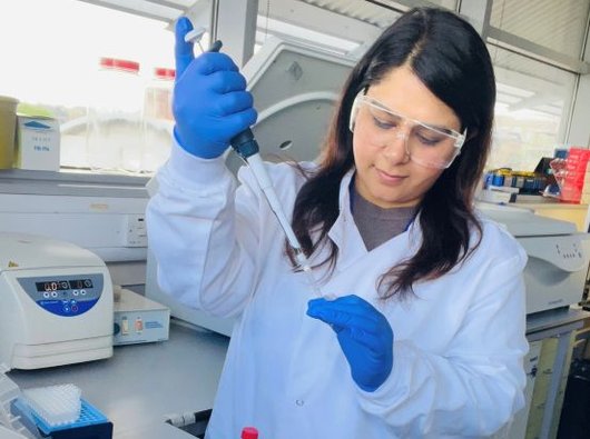 A lady working in the lab wearing lab goggles and a white lab coat, holding a pipette.
