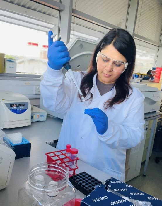 A lady working in the lab wearing lab goggles and a white lab coat, holding a pipette.