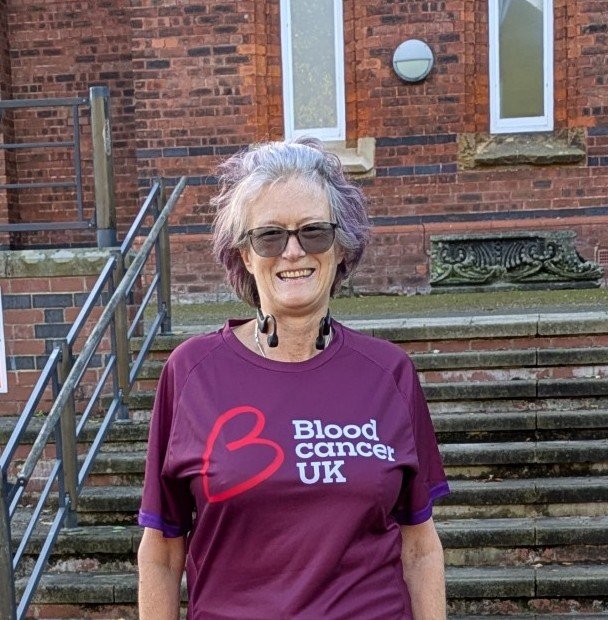 A smiling woman, wearing a Blood Cancer UK running top. She is standing in front of steps.