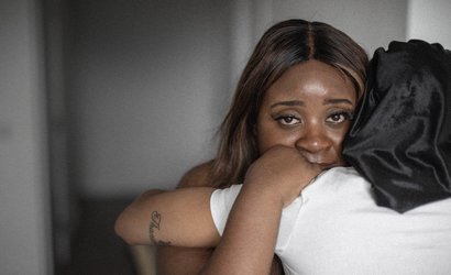A Black woman hugs another woman in a white t-shirt. She's looking directly into the camera.