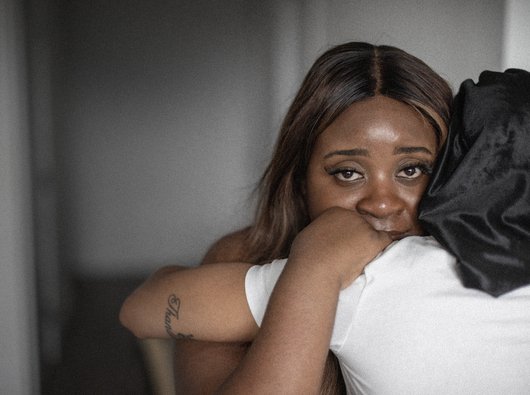 A Black woman hugs another woman in a white t-shirt. She's looking directly into the camera.