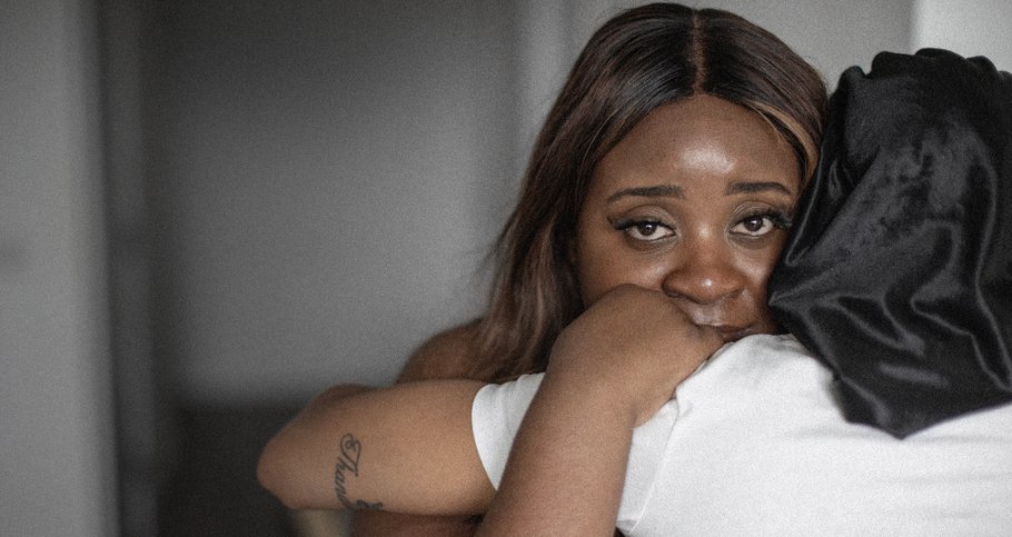 A Black woman hugs another woman in a white t-shirt. She's looking directly into the camera.