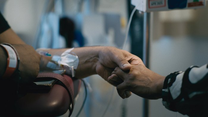 A patient and another person hold hands. The patient has an IV in their arm and a wristband on.