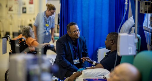A man speaking to a doctor at the bedside. They are in a ward. They are both sitting down.