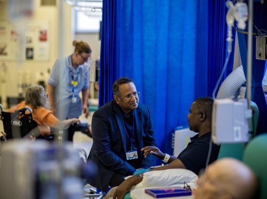 A man speaking to a doctor at the bedside. They are in a ward. They are both sitting down.