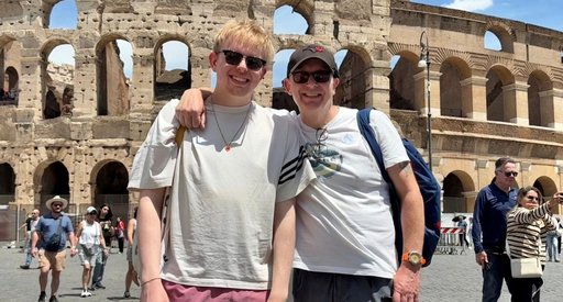 Mark stands in front of the colosseum in Rome facing the camera. He has a hat and sunglasses on. His teenage son stands next to him. They are both smiling.