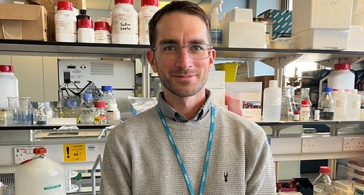 Image of a man stood in a science lab wearing a grey jumper wearing a lanyard round his neck.
