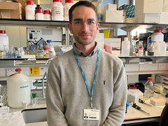 Image of a man stood in a science lab wearing a grey jumper wearing a lanyard round his neck.