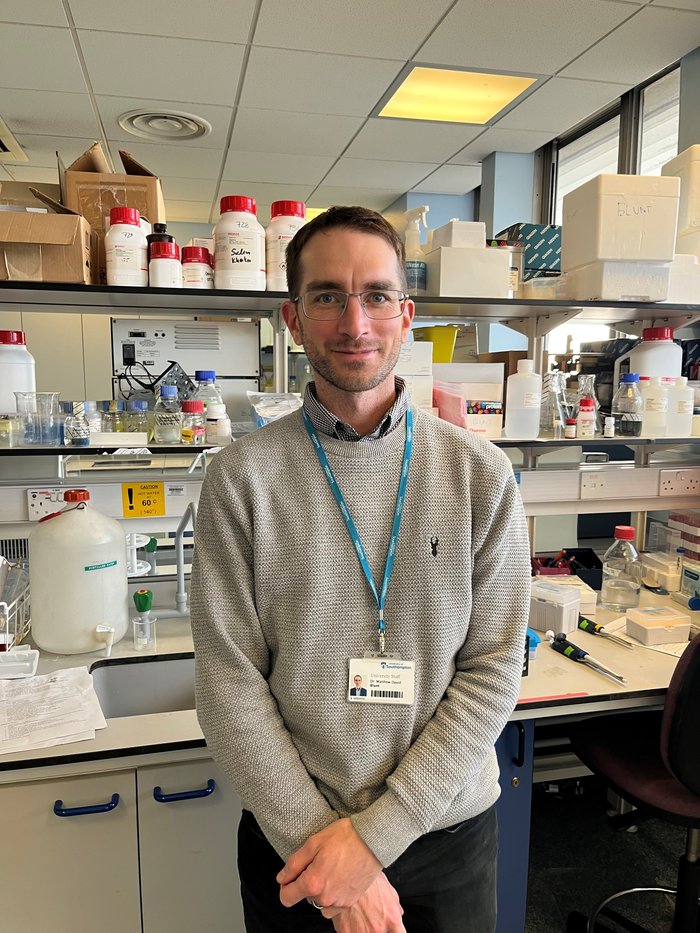 Image of a man stood in a science lab wearing a grey jumper wearing a lanyard round his neck.