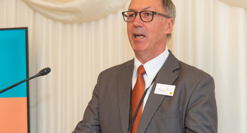 Picture of a man in a suit holding a microphone, speaking at an event.