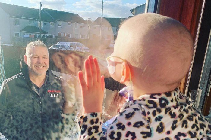 A child undergroing treatment leans up against a window as a man smiled to them from outside.