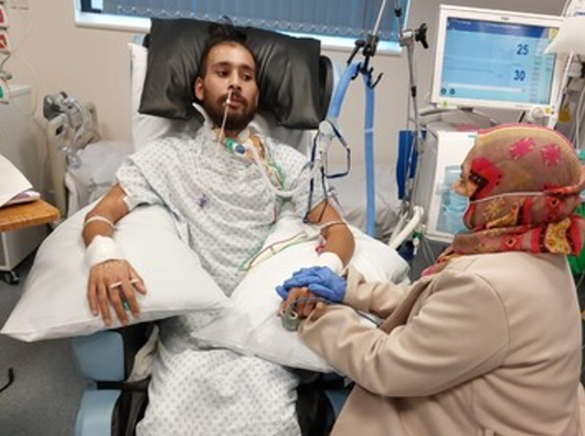 A man is in hospital surrounded by machines and wires. A female relative holds his hand.