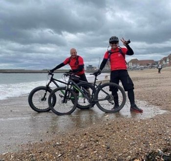 Dean Smith and his friend stand on a stoney beach with their bikes. A grey sky thick with clouds looms behind them. They both smile and pose for the camera happily.