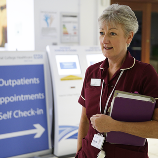A nurse, holding a folder, stands in a hospital waiting room with a receptionist behind her.
