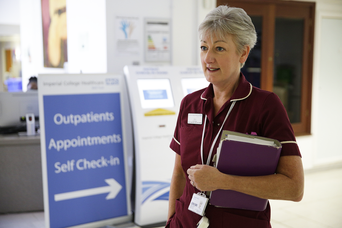 A nurse, holding a folder, stands in a hospital waiting room with a receptionist behind her.