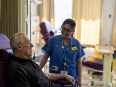 A man sitting in a high-backed chair in hospital, talking to a nurse in blue hospital scrubs standing next to him.