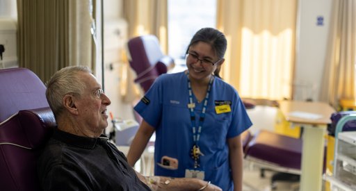 A man sitting in a high-backed chair in hospital, talking to a nurse in blue hospital scrubs standing next to him.