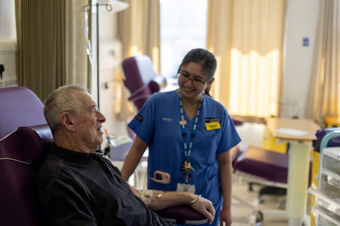 A man sitting in a high-backed chair in hospital, talking to a nurse in blue hospital scrubs standing next to him.