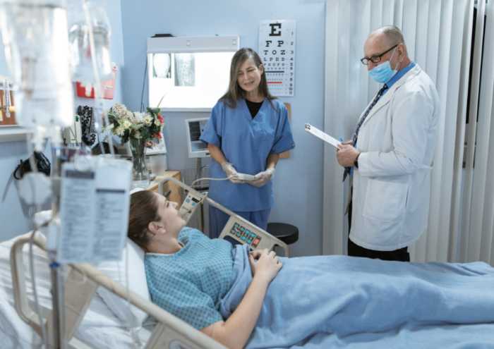 A masked doctor studies a patients chart as he stands over her bed. A nurse also stands nearby.