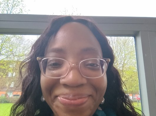 Precious, a Black lady, stands in front of a window and smiles at the camera.
