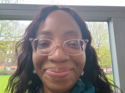 Precious, a Black lady, stands in front of a window and smiles at the camera.