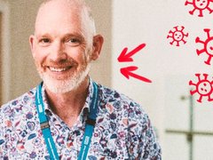 Image of a man with a grey beard and brown eyes, wearing a blue NHS lanyard and colourful shirt smiling at the camera. Hand-drawn red blood cells in the top right hand corner.