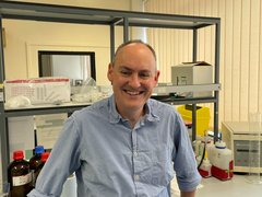 A picture of a man stood in the lab smiling wearing a blue and white checked shirt.