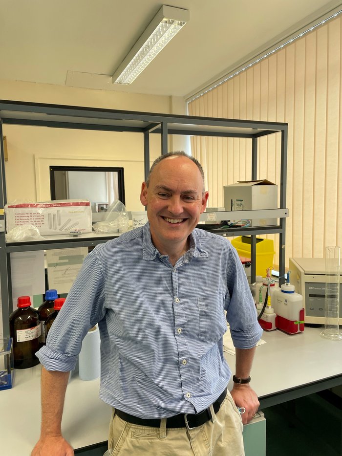 A picture of a man stood in the lab smiling wearing a blue and white checked shirt.