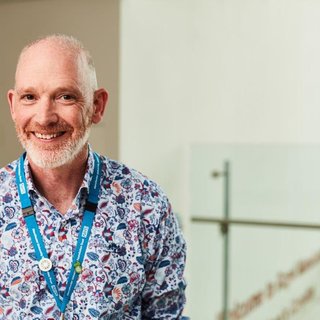 A man stood smiling at the camera wearing a patterned shirt and a blue NHS lanyard.