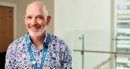 A man stood smiling at the camera wearing a patterned shirt and a blue NHS lanyard.