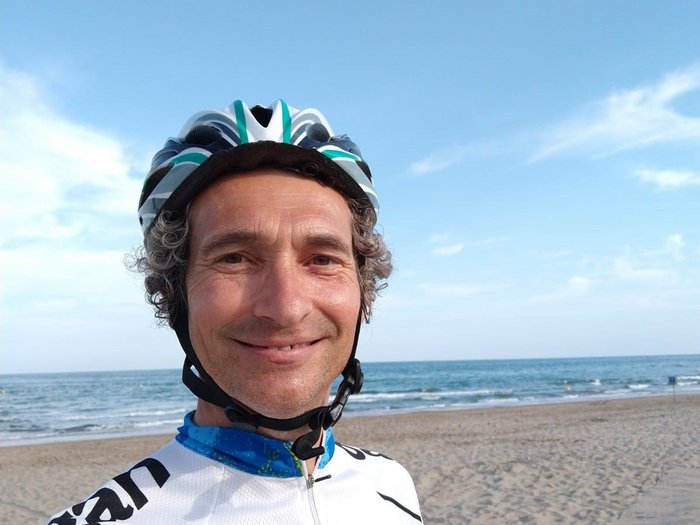 Quentin, who lives with acute myeloid leukaemia (AML), stands on a sandy beach wearing cycling clothes and a helmet.