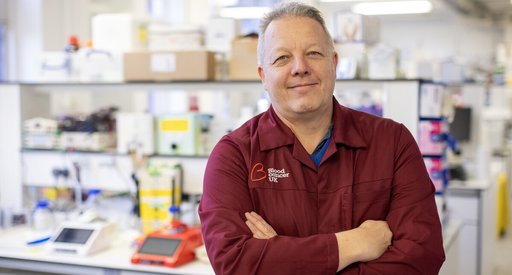 A man stood in a science lab with his arms folded wearing a red Blood Cancer UK lab coat.