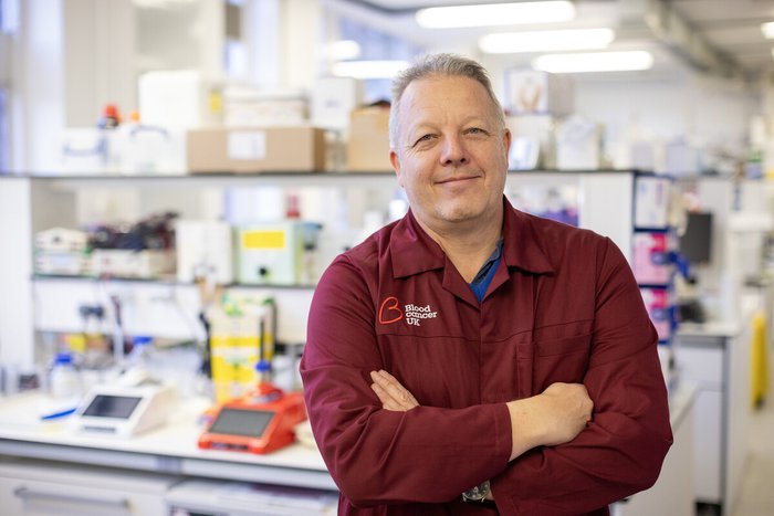 A man stood in a science lab with his arms folded wearing a red Blood Cancer UK lab coat.
