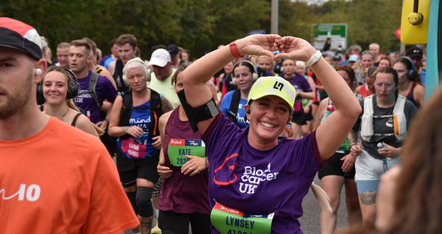 A group of runners takes part in a regional run. In the foreground, a smiling runner in a purple Blood Cancer UK shirt and bright yellow cap raises her arms overhead, while other runners fill the road behind her.